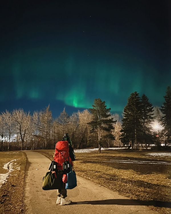 A person wearing a large red backpack stands under the northern lights on a path in a lightly wooded area. 