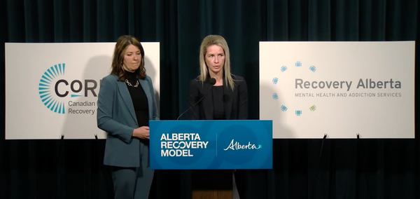 Two women in formal professional attire speak in front of a podium that says "Alberta Recovery Model", with signs behind saying CoRE and Recovery Alberta.