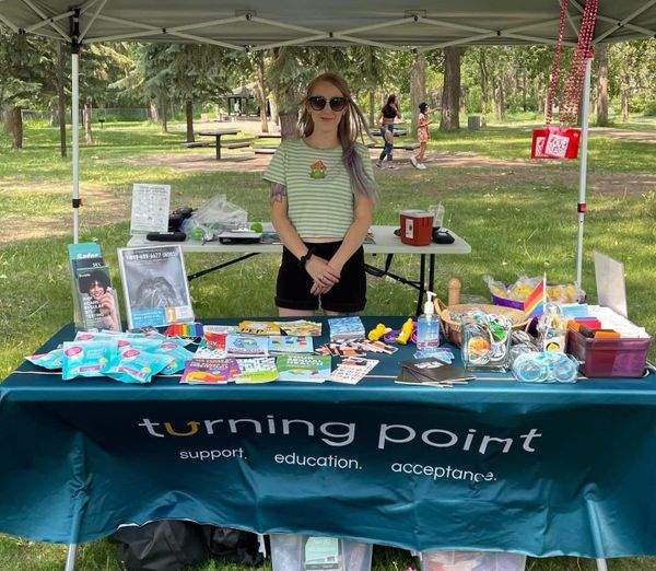 A woman in a green and white striped t-shirt and long lightly purple-coloured hair smiles standing at a table with harm reduction supplies.