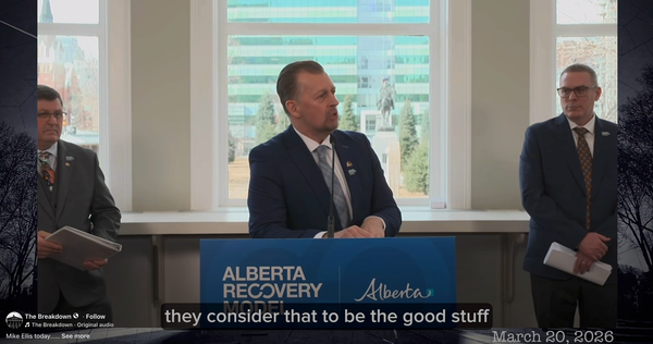 A man in a suit leans on a podium that says "Alberta Recovery Model" while speaking toward someone at the side.