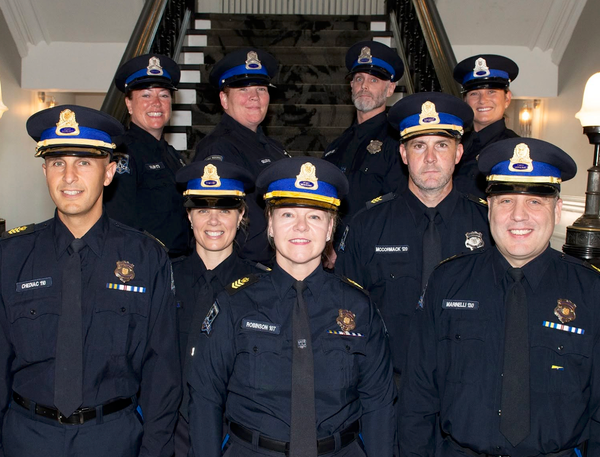Nine police officers in police caps and uniforms smiling in front of an old staircase.