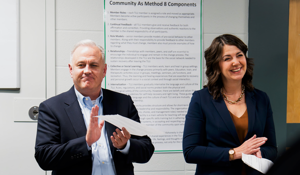A man and a woman, both in suits, clap and smile in a cinder block room inside a jail. 