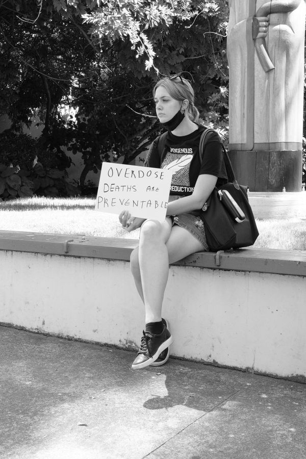 A person in shorts, sneakers and a Indigenous Harm Reduction t-shift holds a sign reading "Overdose Deaths Are Preventable."