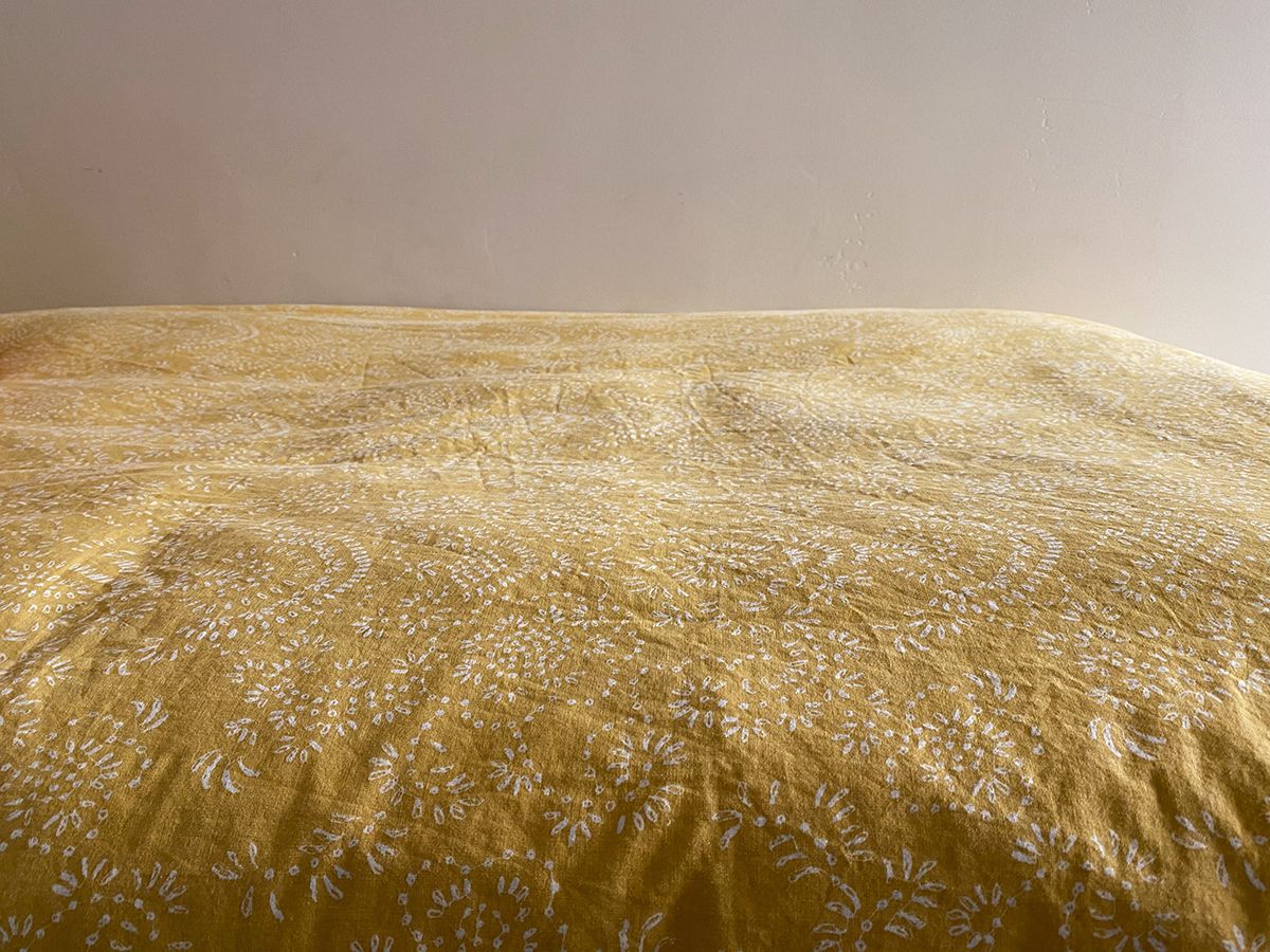 A lightly-wrinkled gold-ish/yellow-ish bedspread with a faint white floral pattern, in front of a blank wall.