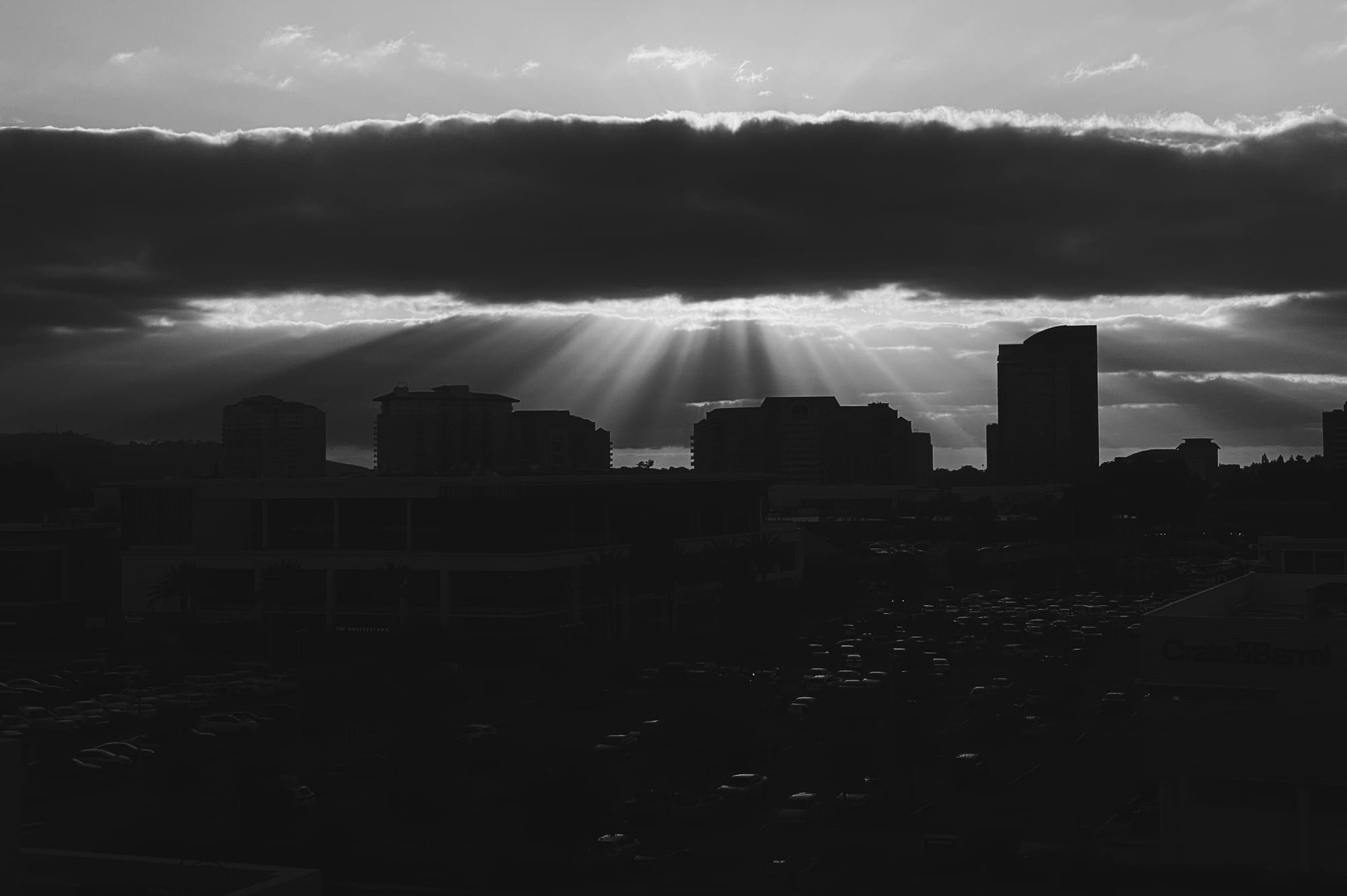 A high-contrast black-and-white photo of sunbeams shining from behind clouds onto a dark city skyline.