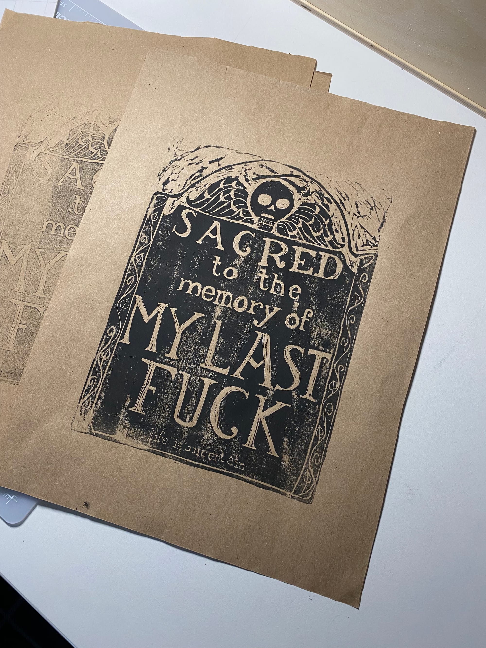A linoleum print in black ink on brown craft paper, the image is in the style of an early New England tombstone with a death's head and the words "Sacred to the memory of my last fuck" and "Life is uncertain" at the bottom