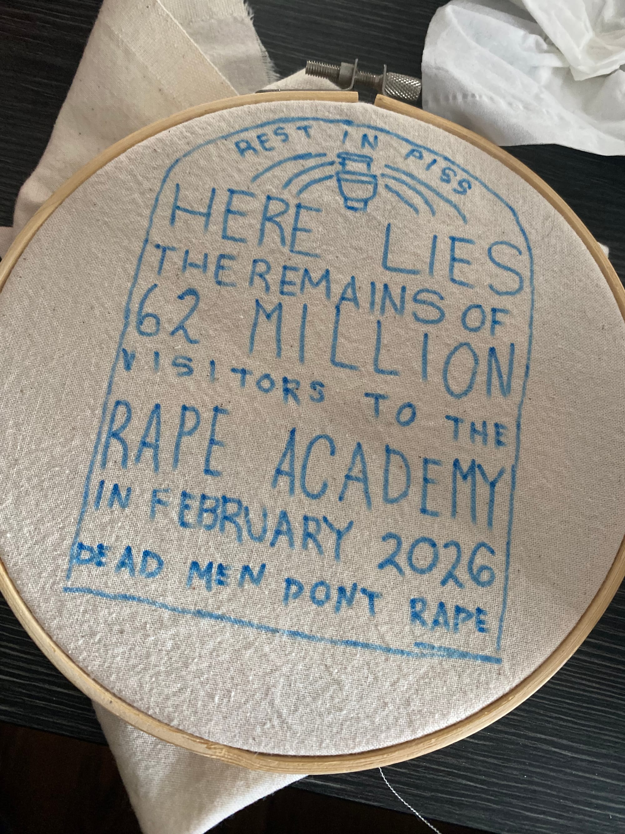 An embroidery hoop fitted with unbleached cotton, with a design in blue marker, resembling a tombstone that says "Rest in piss," "Here lies the remains of 62 million visitors to the rape academy in February 2026" and "Dead men don't rape"