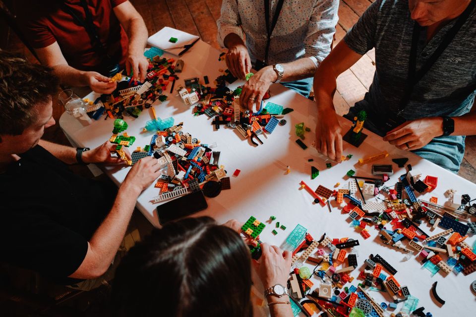 People around a table with lego pieces scattered around, there are five people creating small models