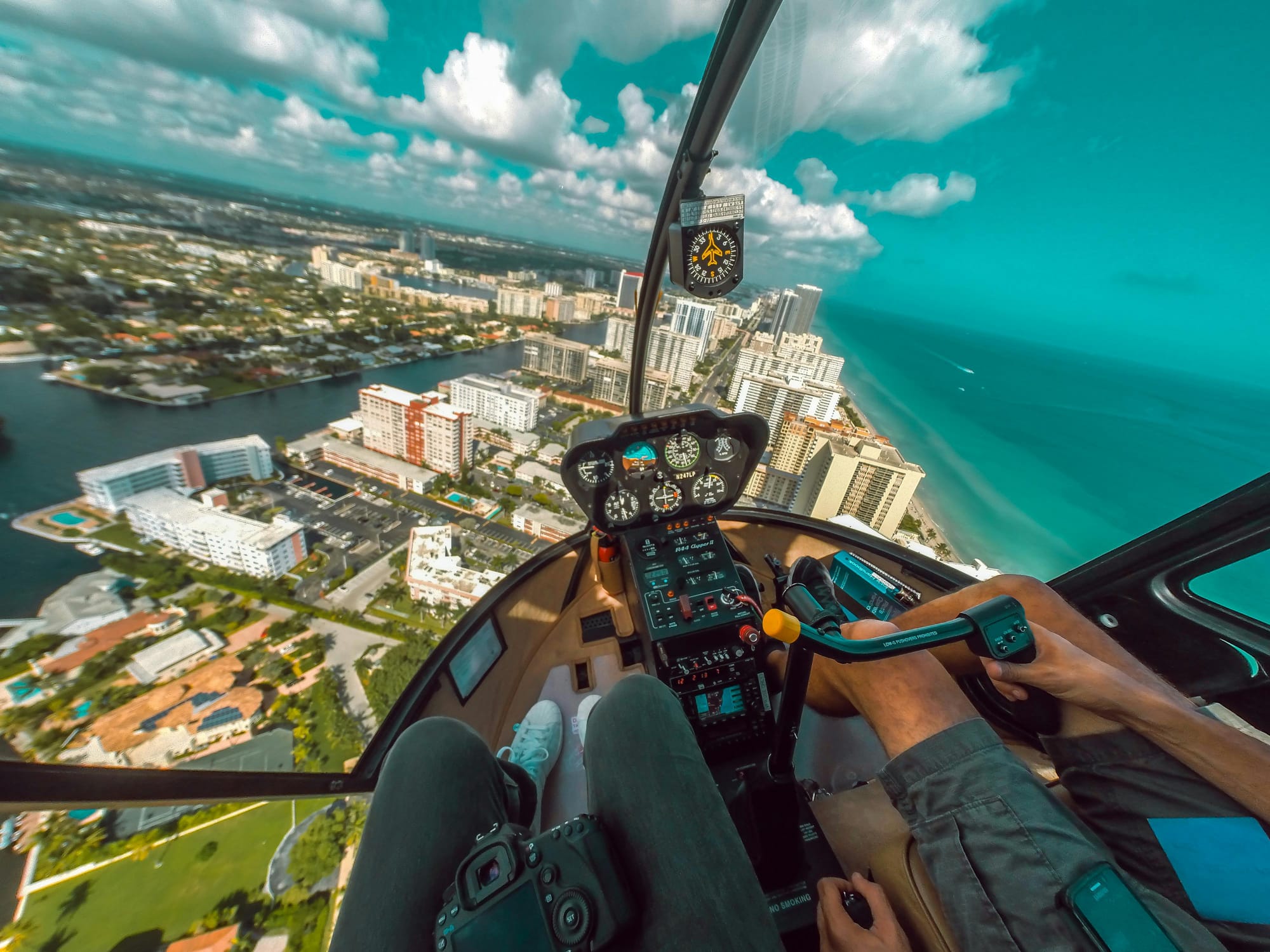 View of a coastal city from the cockpit of a Robinson R44 helicopter. The legs of the front seat occupants are visible. The passenger has a camera on their lap. The pilot is balancing a mobile telephone on his thigh. 