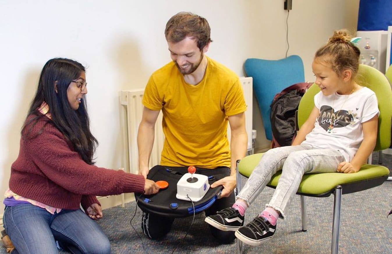 Woman in maroon sweater configures adaptive games controller with smiling man in yellow shirt. Young girl sits in green chair next to them, smiling.