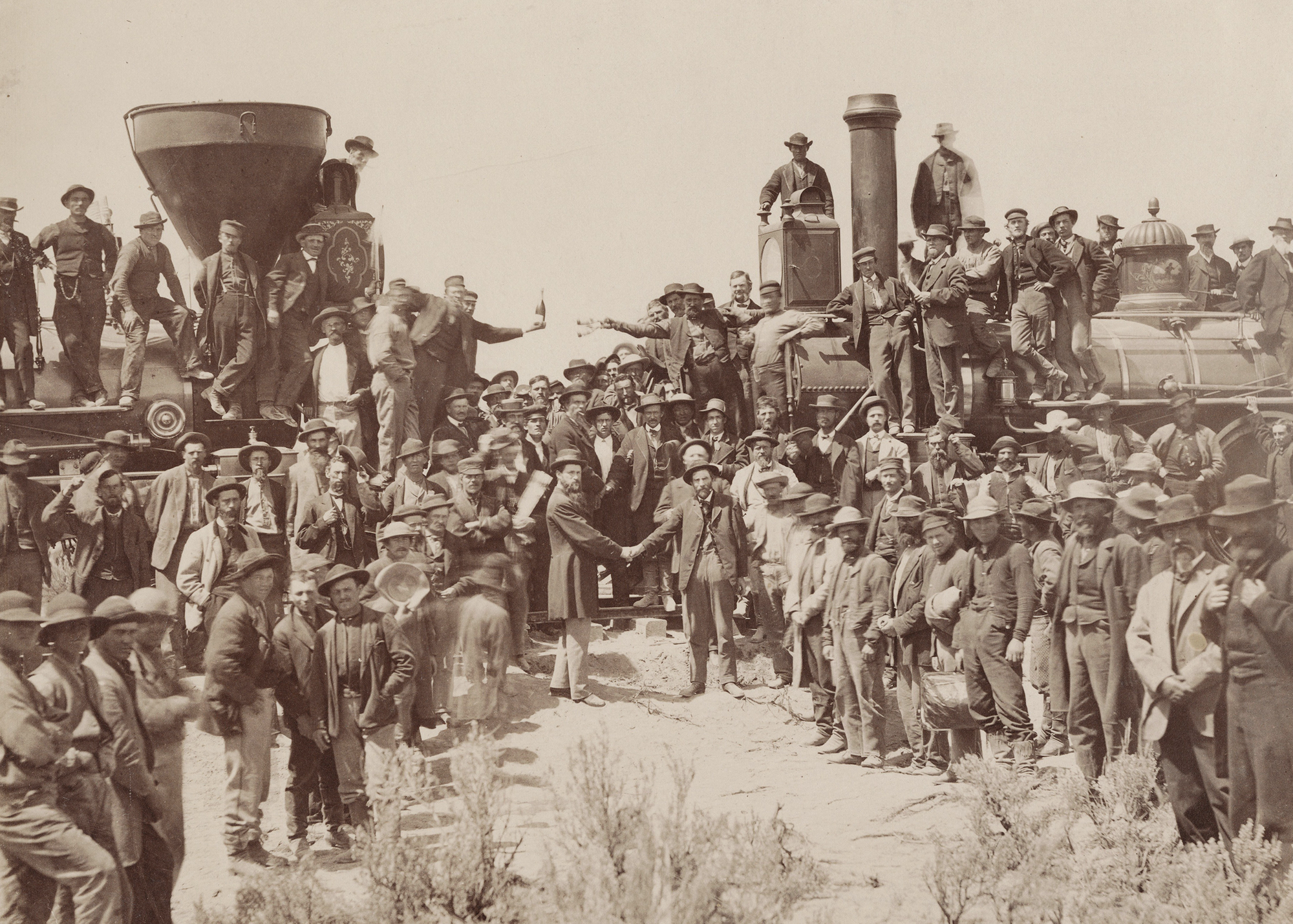 Historic black-and-white photo showing a large group of men celebrating the joining of two steam locomotives. 