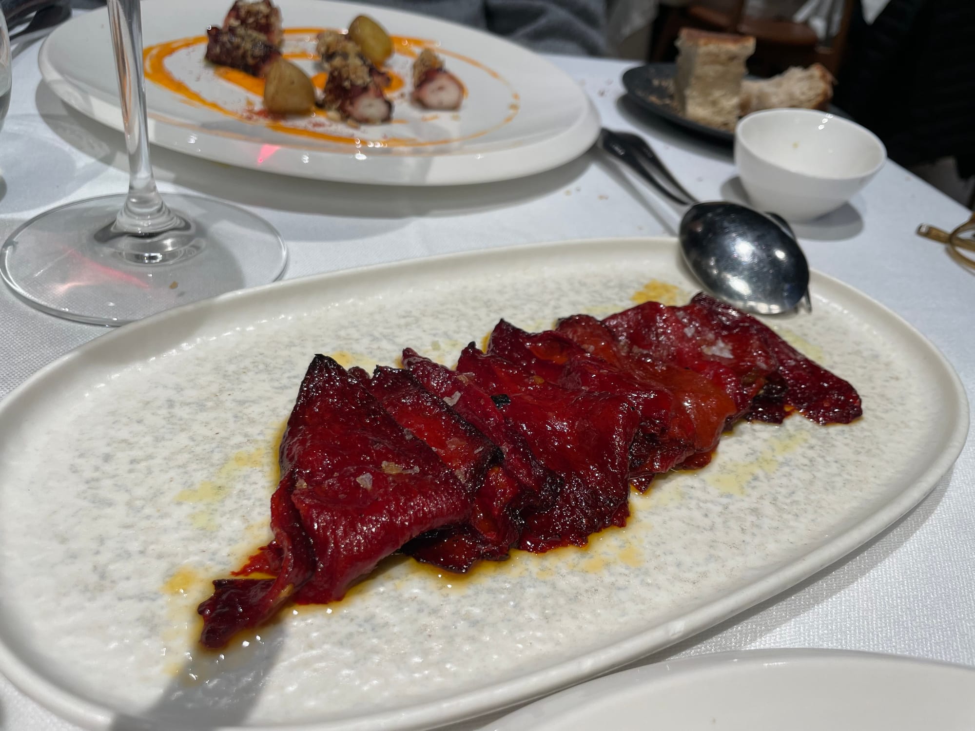 dark, richly caramelized slices of red pepper on a pale grey oval stoneware plate, with other luscious tidbits and wineglasses in the background