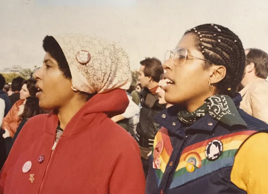 Two young Black women at a 1979 march for gay rights in Washington, in colorful gear, protest buttons and bandannas, seemingly absorbed in a speech being given offscreen