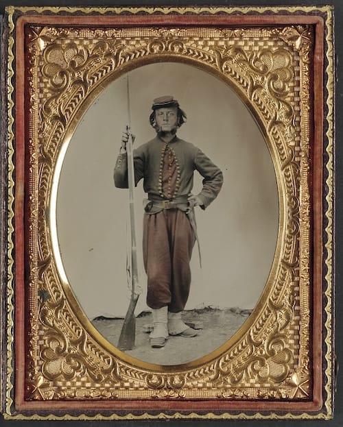 Soldier stands with his hand on his hip propping up a rifle with a bayonet. He has a stoic expression and also mutton chops.