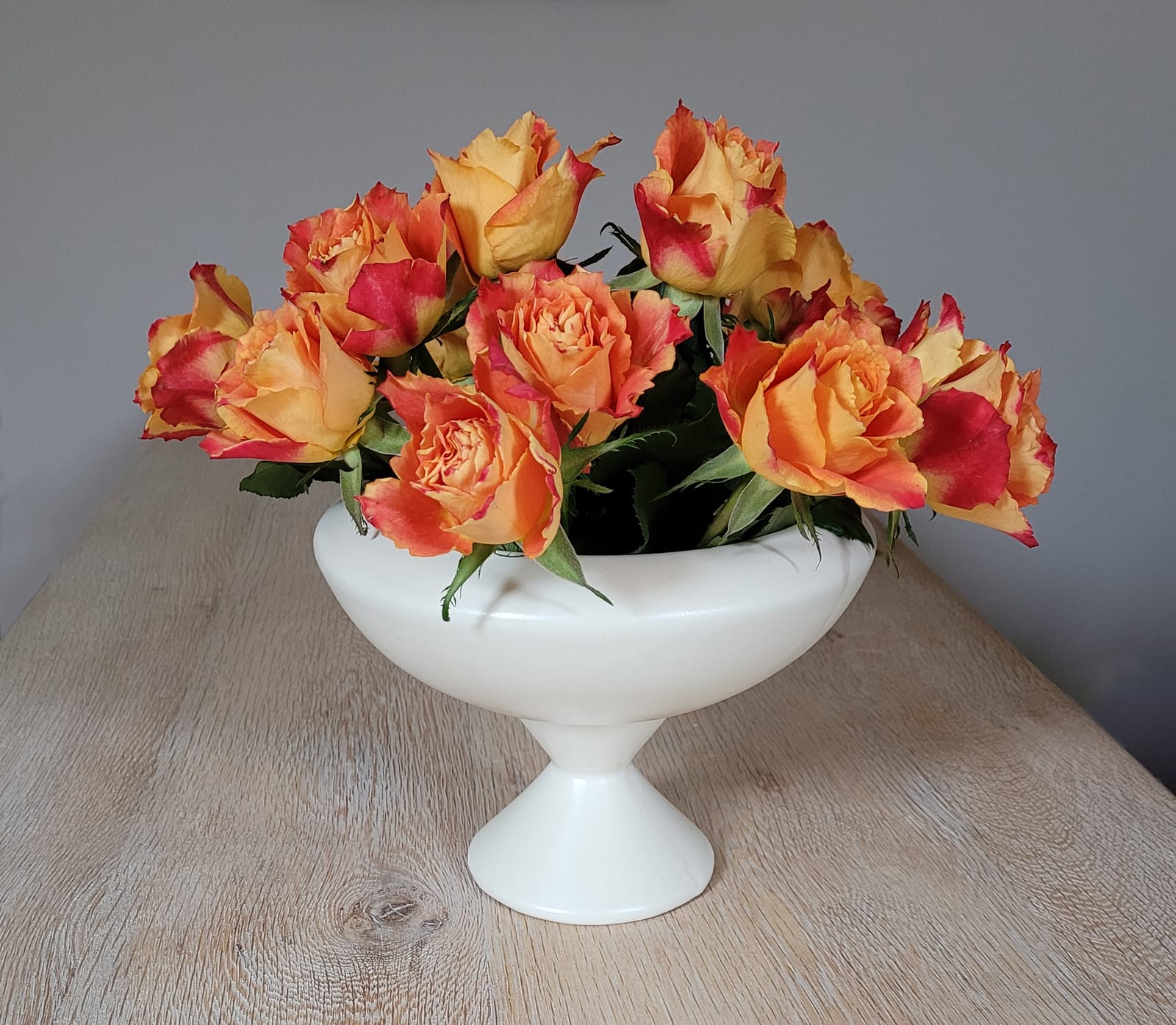 A cream ceramic chalice holding vivid orange-crimson roses with their foliage