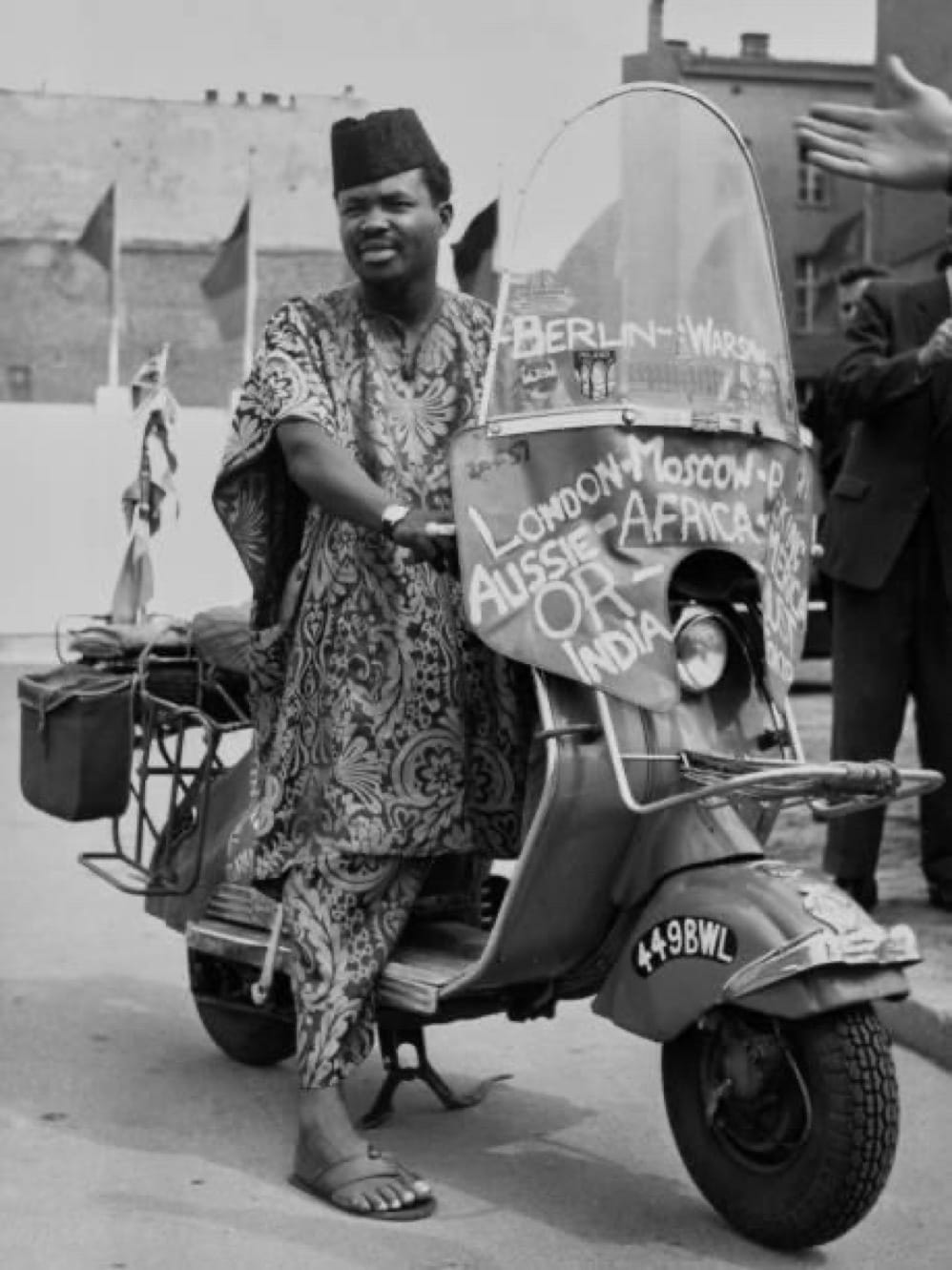 Black and white photo of Ọlábísí Àjàlá posing on his scooter; he wears a traditional African smock in an intricate print, a dark cap, and leather sandals