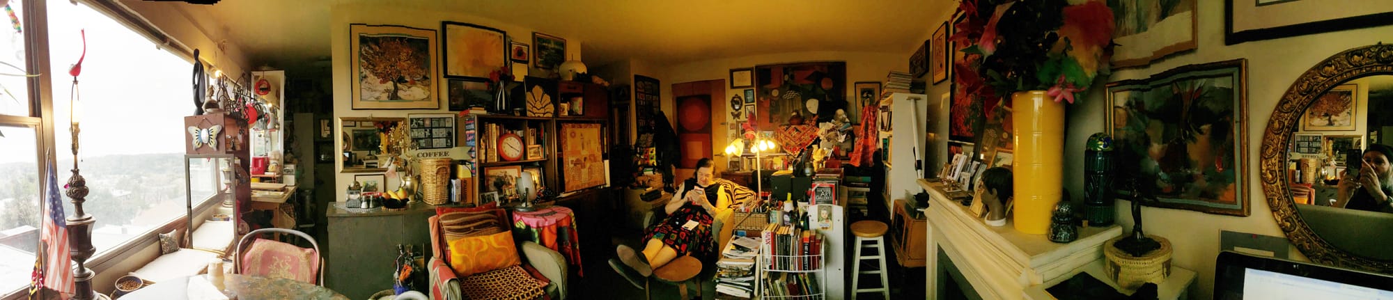 [ALT: L-R: A wide, curtainless, shadeless picture window with details of the exterior blown out by sunlight. Inside, small American and Maryland flags, a lamp without a lampshade with a bare bulb, illuminated, and a bird sculpture on the finial, a dish of dry cat food, a cat bed, a tiny pillow, a chair with some old ornate fabric on it, a piece of butterfly art with a mirror in the middle of it, a mirror, a shelf with assorted kitchen stuff on, a small modern-art-looking figurative sculpture, a small bird sculpture, another lamp with a bare bulb on, a hanging rack with pots and pans, another mirror, a red toaster, an old can opener, a shadow-darkened area (the tiny galley kitchen), another mirror, a bunch of framed art along the wall, an arrow-shaped sign with COFFEE on it, a diner-style napkin holder with a brass snail on top of it, salt and pepper shakers, a bowl with plastic fruit (banana, orange, pear), a metallic vase with some metallic floral-shaped stuff comingout of it, a basket, some books, a bookshelf with a clock on it, a chair with a pillow on it covered with wacky fabric, another pillow with wacky orange fabric, some wacky checked fabric on the seat, a small table covered with some brightly-colored floral-inspired fabric, a small vintage Vornado fan, a vertical-striped vase with paper flowers in it, a batik-looking wall hanging, my mom Jessica sitting in her chair with her feet up on asmall wooden stool looking at a notebook, a ceramic bird sculpture that opens and you can keep tiny junk in it, a fabric sort of Rothko-inspired-looking wall hanging on the apartment door, a metal butterfly on the wall near the door, soem tiny framed artworks, a really big artwork taking up a large portion of the wall near the door, a multi-bulb lamp with no shades except for one, and it’s on its way out, a basket with notebooks in it, a can with pens and scissors and stuff it it, some art books on a wire shelf, some fabric with tiny mirror sewed into it, a bunch of postcards, the top of a Picasso, a bookshelf, full, another wall full of framed art, an off-white painted mantel, framed photos on top the mantel, a small plastic figure of a boy’s head, a big yellow ceramic vase with paper and plastic flowers in it, an old glass insulator that got decorated with some metallic markers, a glass vase with a glass ball in it, a small basket with a metal figurine on top of its lid, a computer monitor, another mirror with me in it taking the picture.