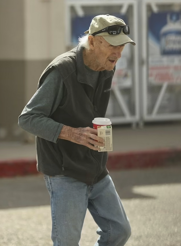 Actor Gene Hackman, age 94, in jeans, a fleece vest, ball cap and big sunglasses, thin and a little stooped, holding coffee and a boxed pie in his right hand