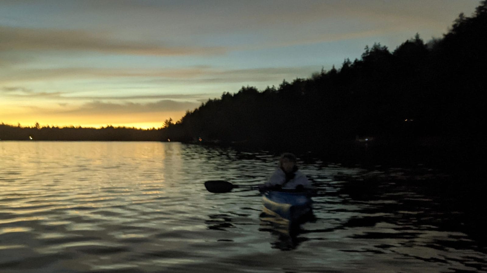 The author dimly visible in a kayak during the April 8 eclipse, just before the totality; light the color of brass on the water, a sliver of lemon above the treeline, and above it a sky full of eerie blues, mauves and orange-grays