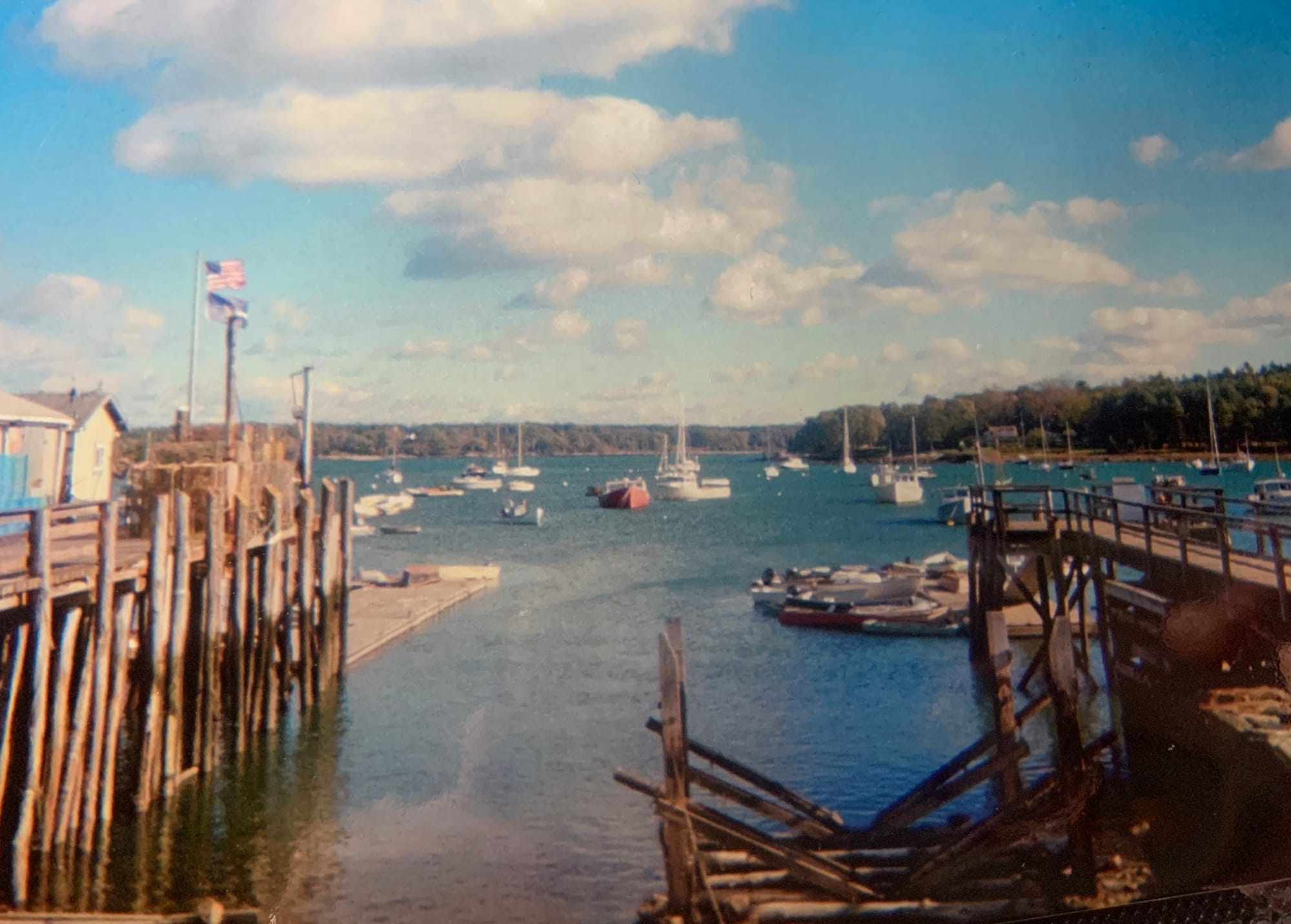 Seaside view of a small harbor in Maine, sailboats at anchor, old wooden piers, forest, clouds, pale blue sky