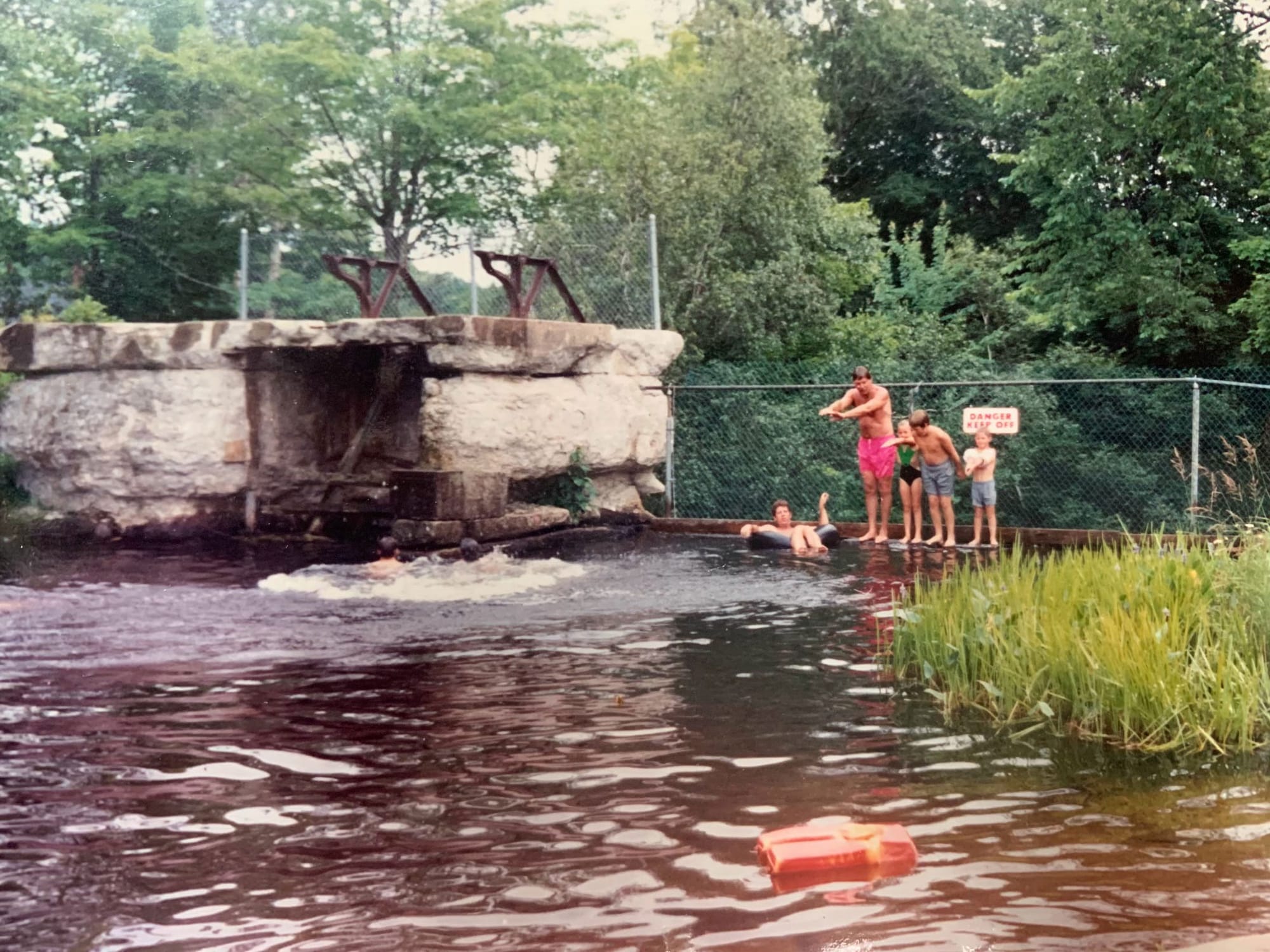 A dad teaching three little kids in bathing gear to dive in a pond. They're standing next to a wire fence with red lettering (DANGER KEEP OFF); a son lolling in an inner tube nearby