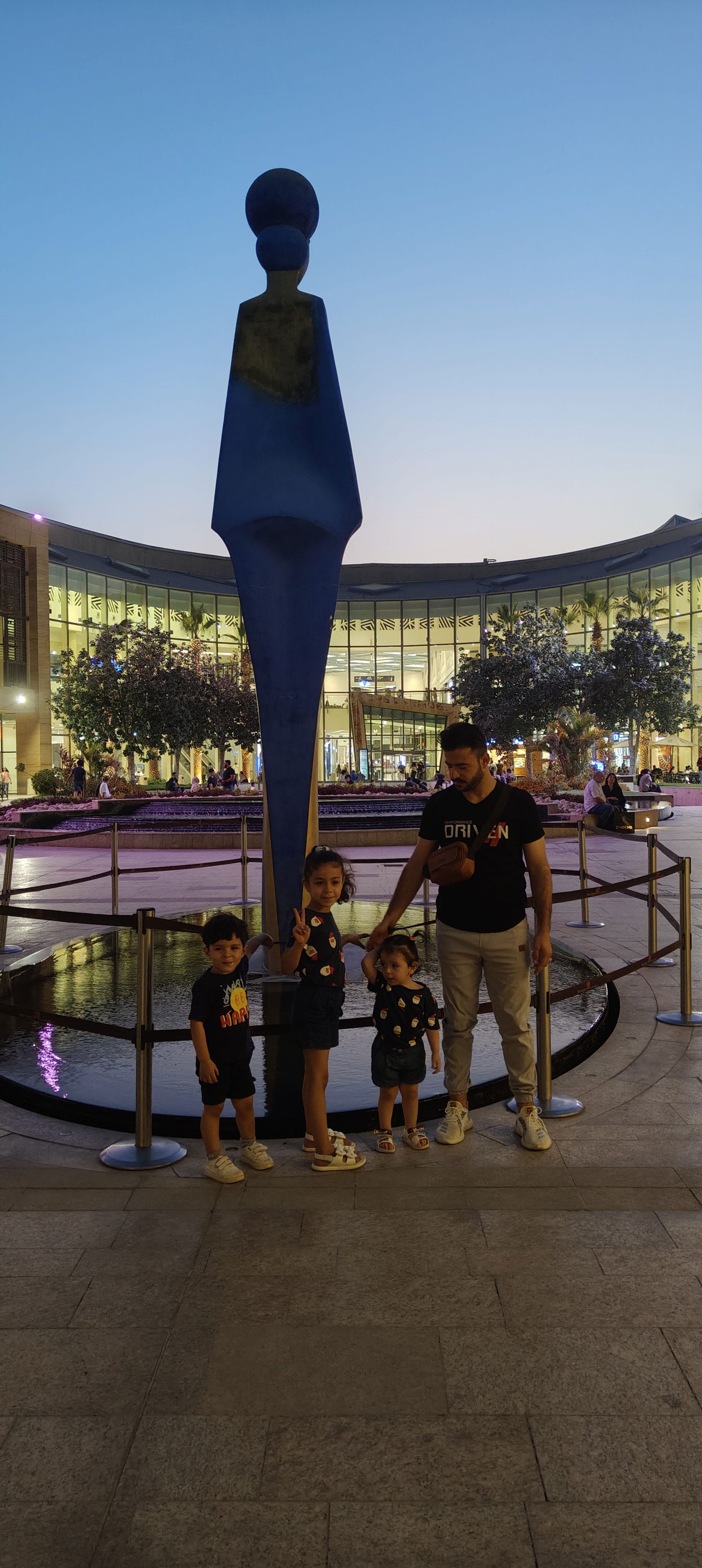 The author and his children on a walk in Egypt, standing before a fountain and a tall sculpture at sundown; behind them, a large well-lit building
