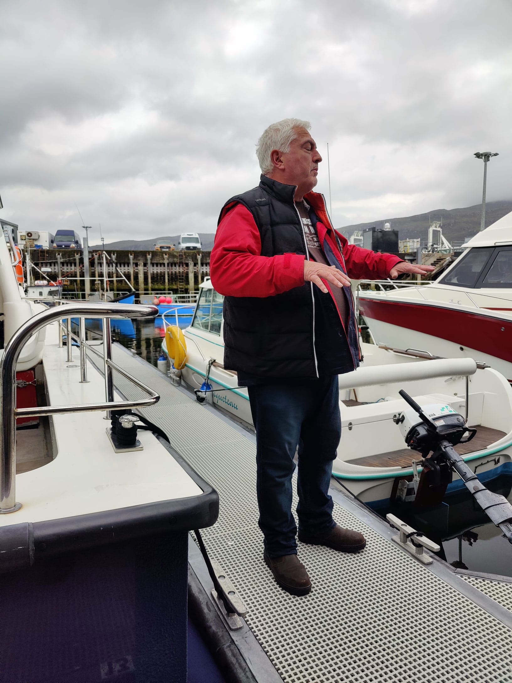 Noel, holding forth on the dock in a light weatherproof jacket with red sleeves, a puffer vest and a dignified expression, in Ullapool Harbor