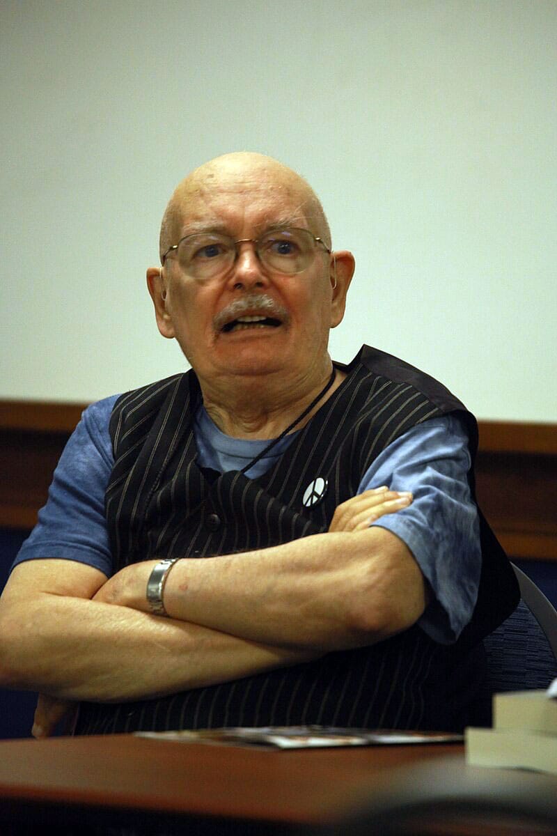 Denny O'Neil, bald, lively, bespectacled, in a striped vest and peace sign badge