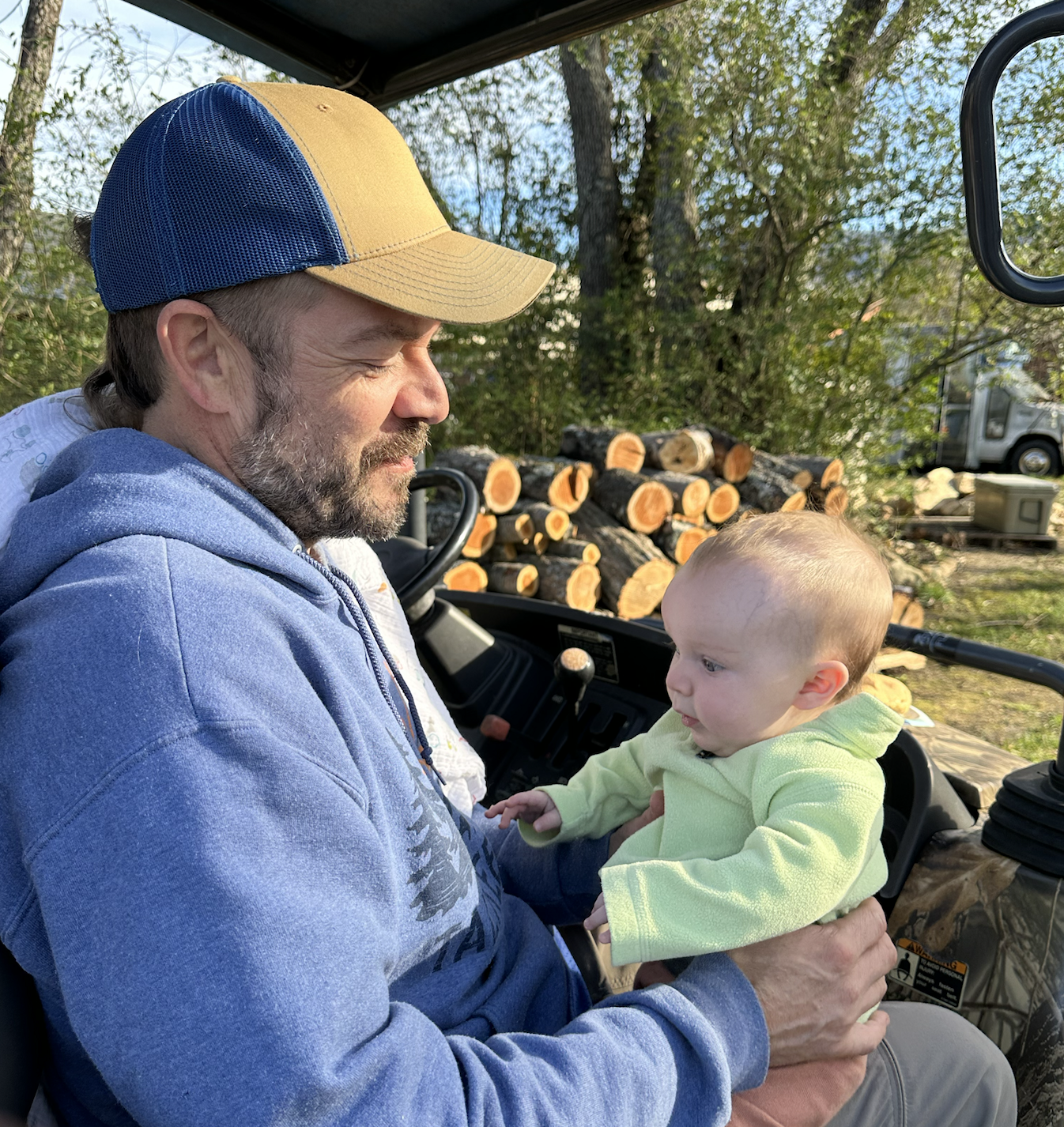 A white man sits in a tractor cab holding an adorable small baby. The man is scruffy and wears a yellow-and-blue ballcap. The baby wears a soft green hoodie and is old enough to be sitting up by himself. 