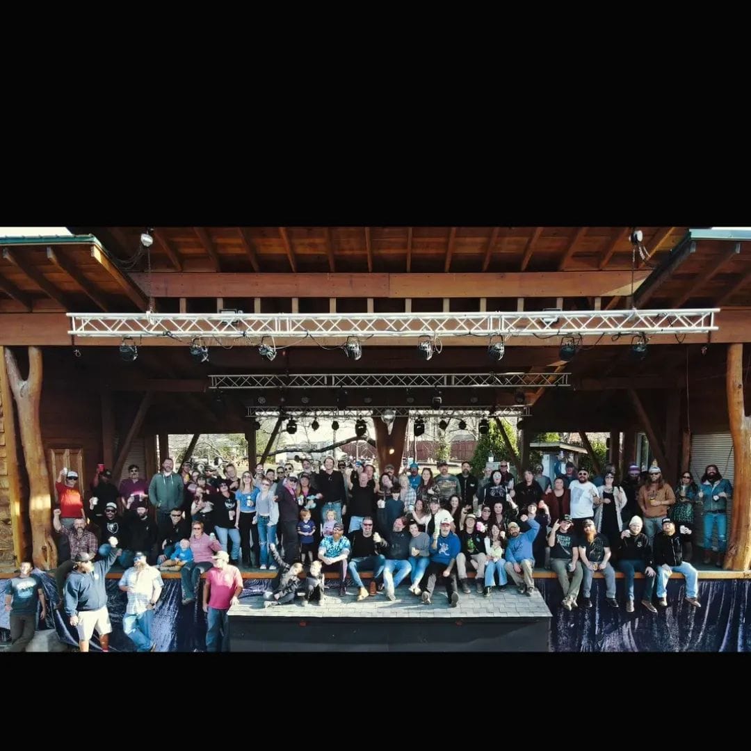 A wide panoramic shot showing an outdoor music stage on which a hundred or so people are posed in a group. In the front row some people are seated or standing against the stage. Many are waving or holding their hands up to the camera. There are several rows of people standing on the stage proper, arrayed in a couple long rows.