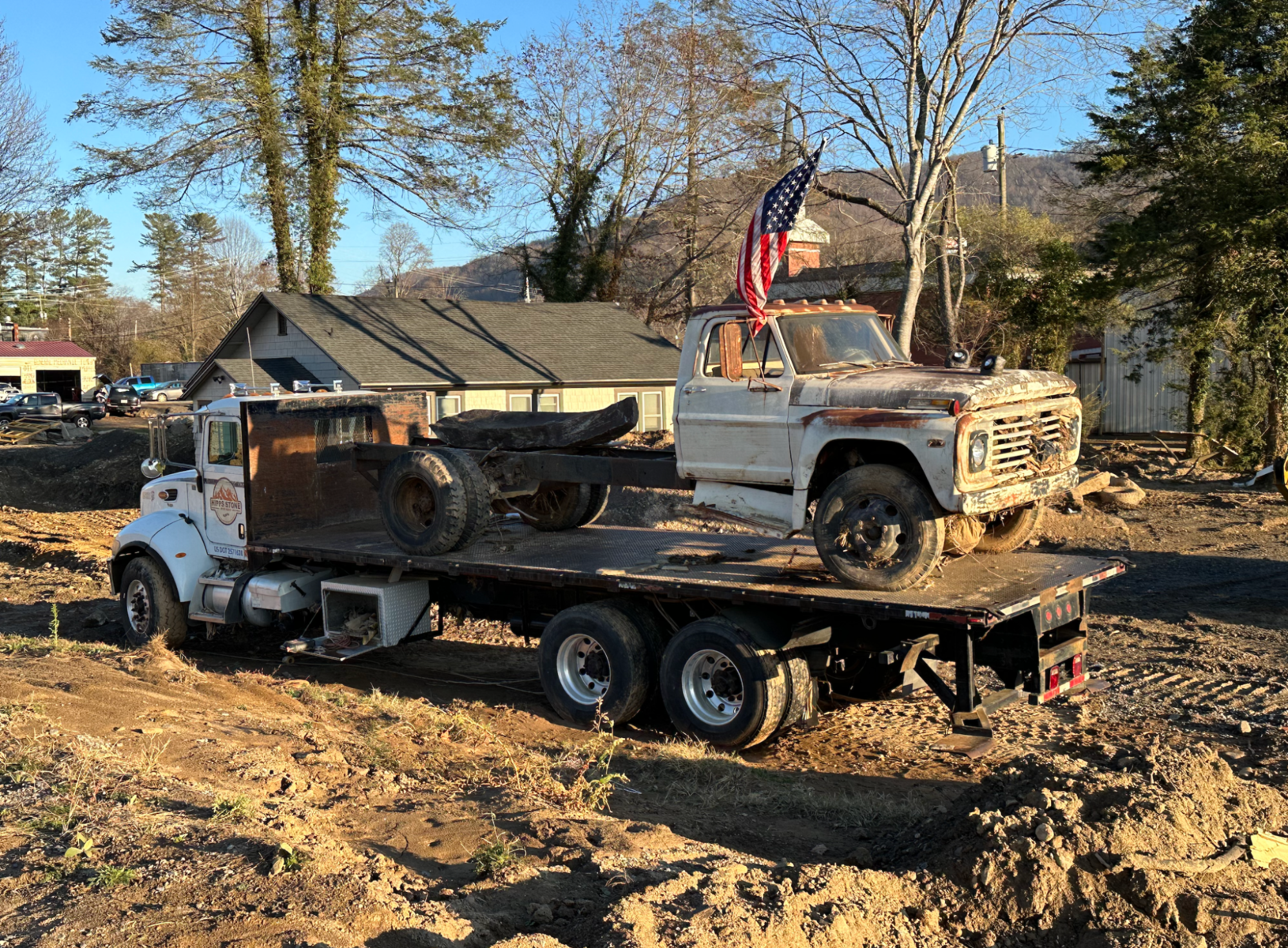 A truck with a white carriage sits in a muddy field. On the bed of the truck is another truck, old and vintage. An American flag hanging from a pole drapes over the older truck’s cab. 