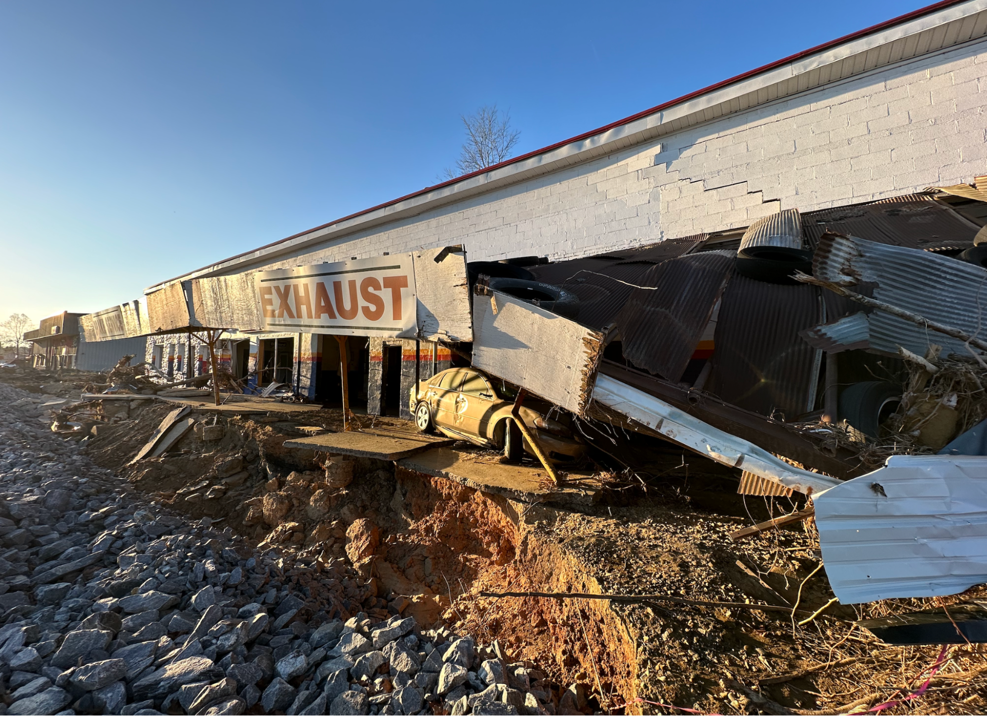 The wreckage of an autoparts and repair store gleams in the afternoon light. You can make out the word “EXHAUST” along the building’s front in big red letters and a crushed car. A corridor of dirt, then of stones, runs along the front of the building. 