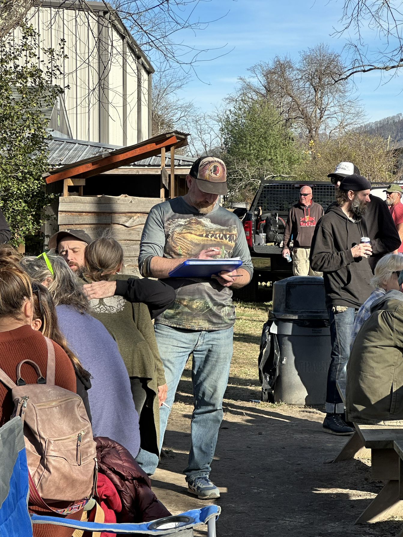 A white man in a grey-and-brown jersey, a ball cap, and jeans writes on a clipboard. On either side of him are two picnic tables with people. At one of them, a man is hugging a woman in a comforting way. 