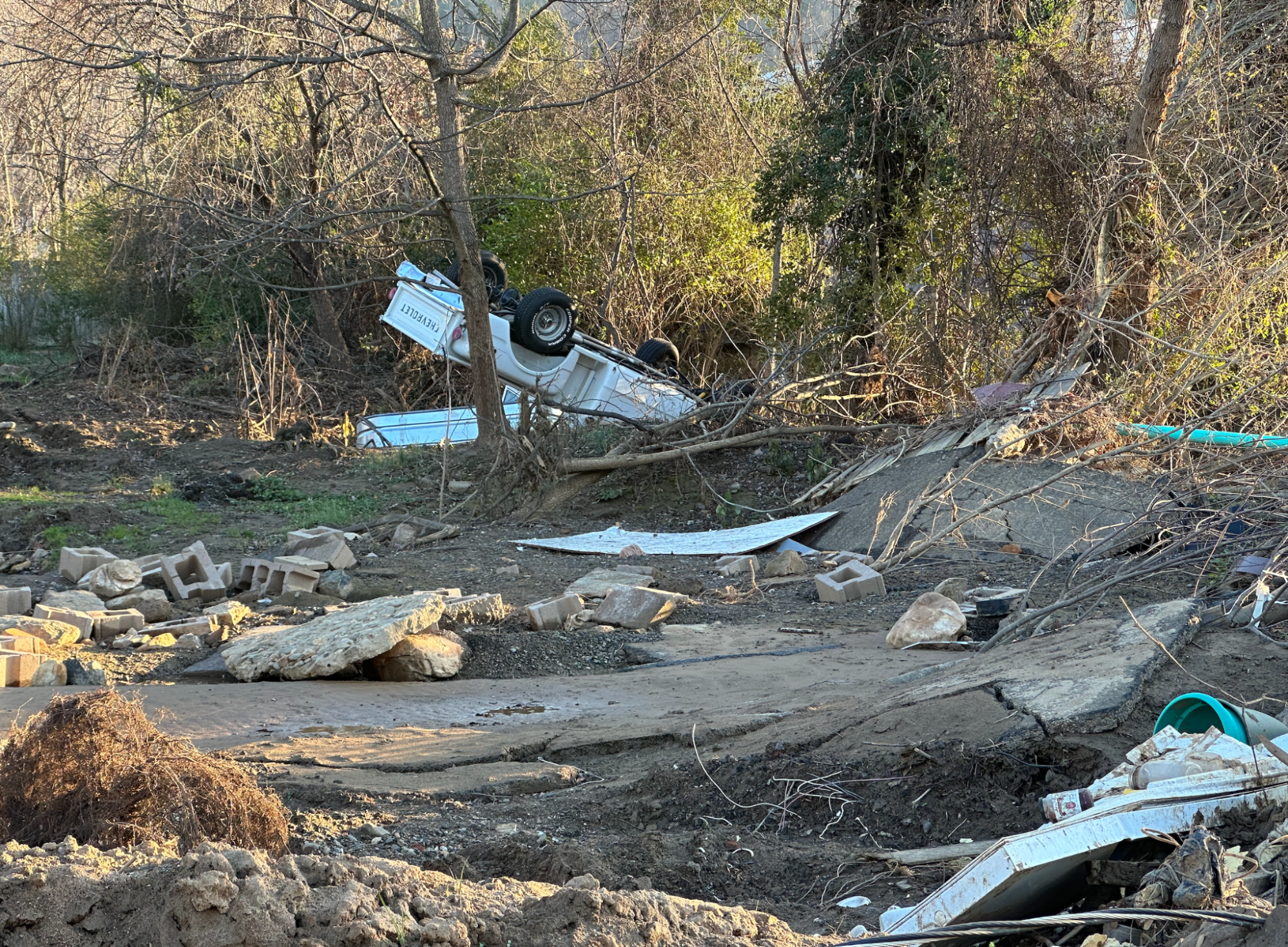 A white truck lies upside down again a backdrop of trees and vines. In the foreground are concrete blocks and great slabs of concrete. 
