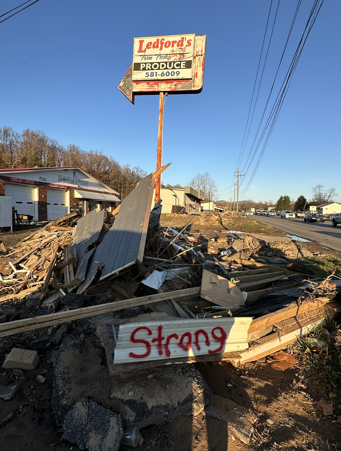 A large pile of building debris sits along the highway. A dilapidated old sign for Ledford Produce rises nearby. Someone has spraypainted “STRONG” in red letters on one of the pieces of debris. 