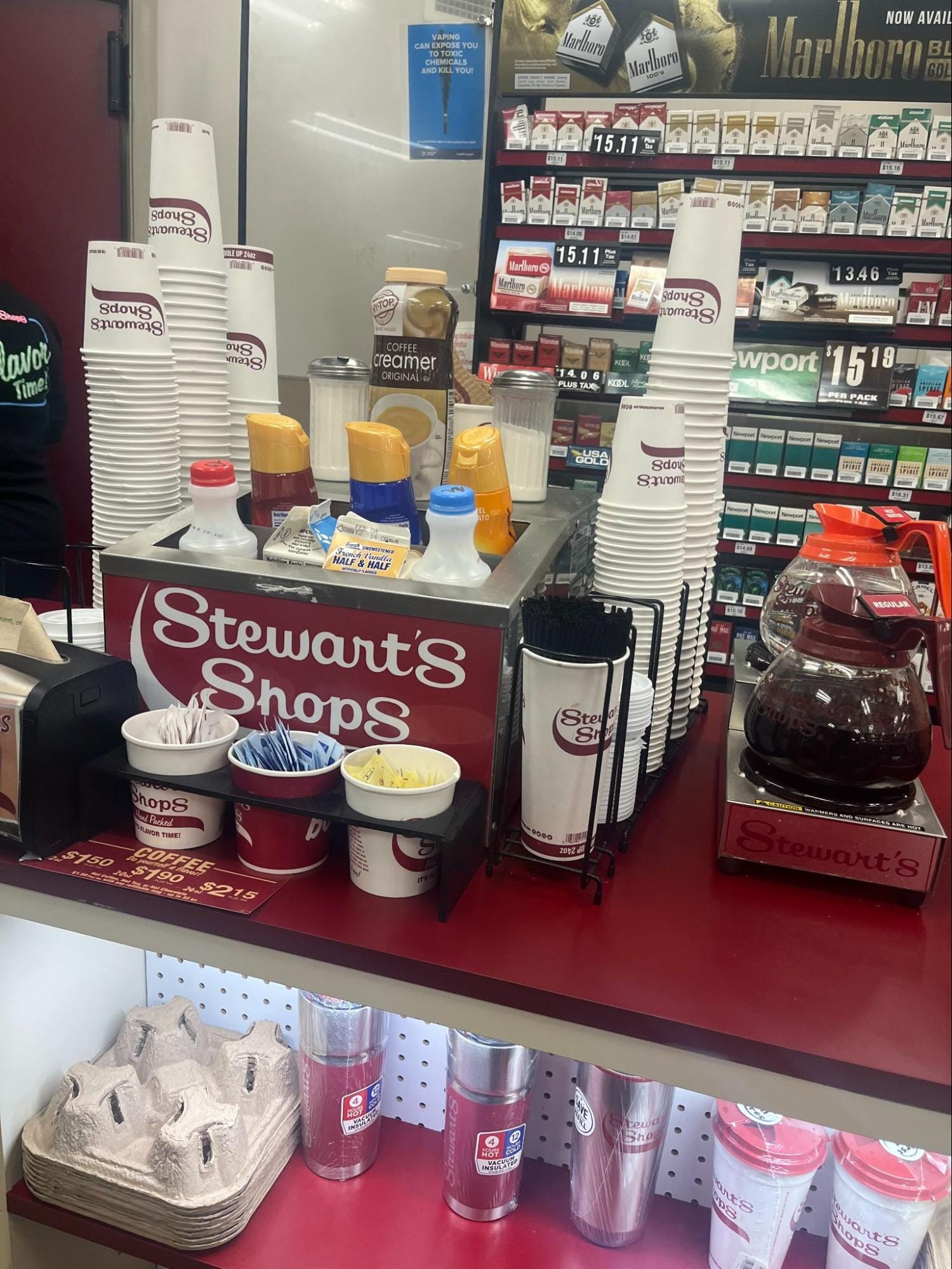 A gas station counter featuring self-service coffeeposts, stirrers and creamers; on the wall behind, a large selection of cigarettes