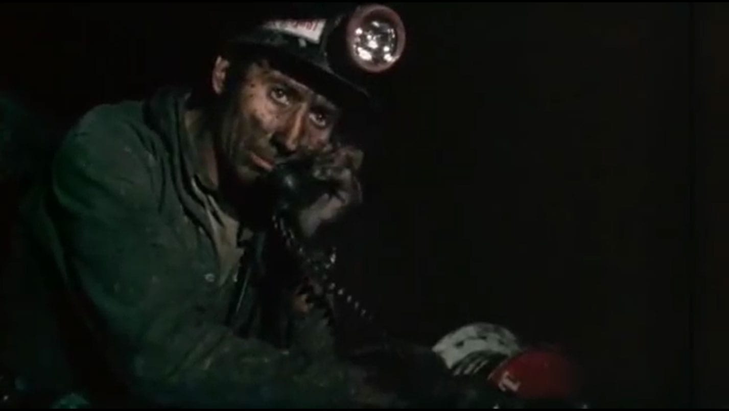 In a screenshot from 'Harlan County, USA' a miner wearing a hard hat with a lamp in the near-darkness of a coal shaft, face blackened with soot, talks on the phone; his expression is worn, worried, and deeply alive