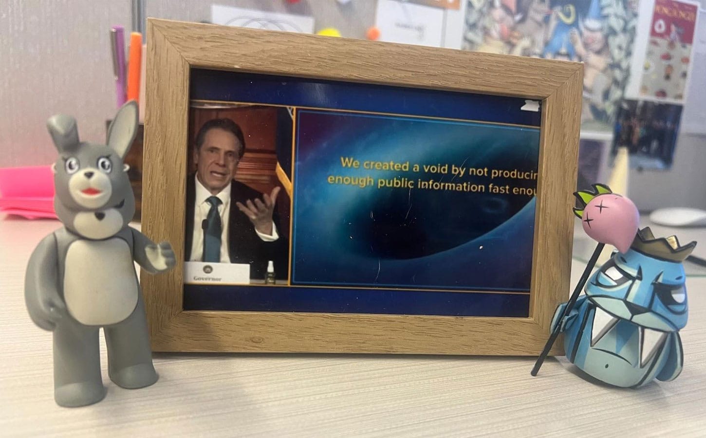 Wood-framed photo of former New York governor Andrew Cuomo, with a quote: 'We created a void by not producing enough public information fast enough.' The frame is on a light wood-grained desk,, flanked by figurines of a teddy bear in a rabbig mask, and a stern-looking blue walrus king holding a pink scepter