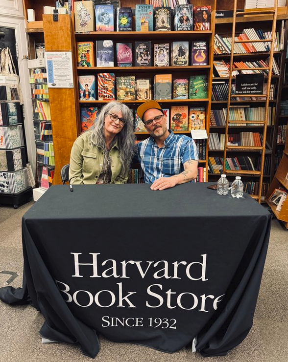 Niko Stratis and Luke O’Neil seated before colorful bookshelves at the Harvard Book Store ('Since 1932')