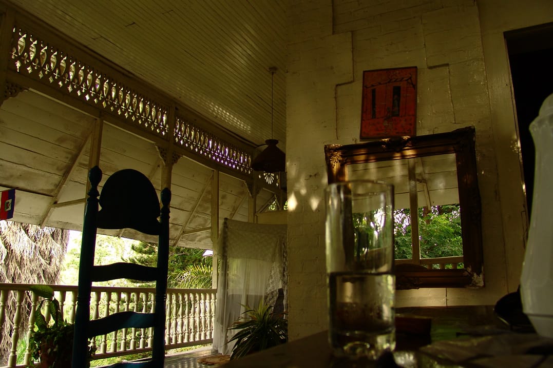 A shade-darkened porch. Visible are a ladderback chair and a half-full glass on a table with a wooden-framed mirror against a side wall