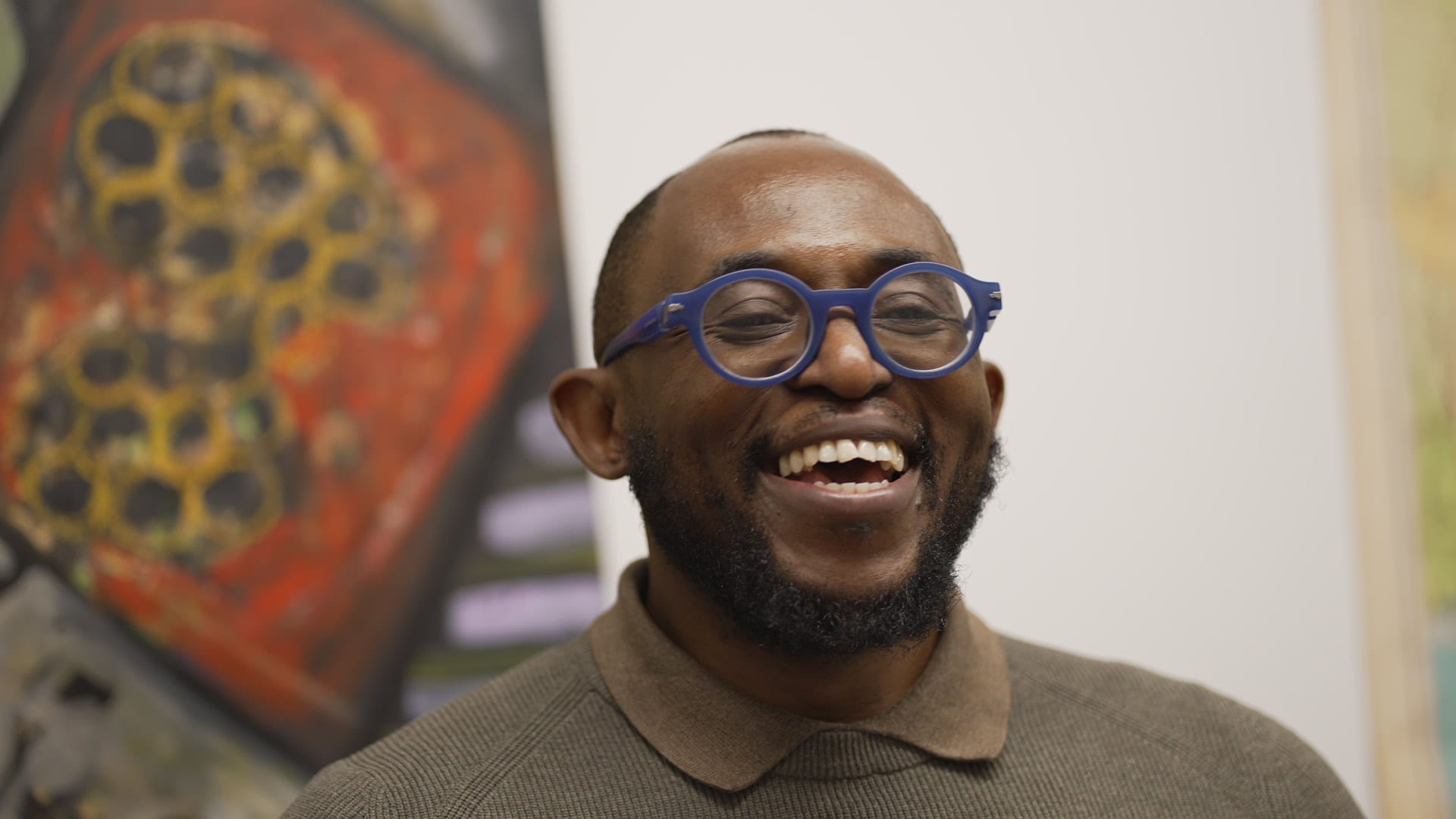 A laughing Black man in snazzy blue glasses and a sweater over a brown polo