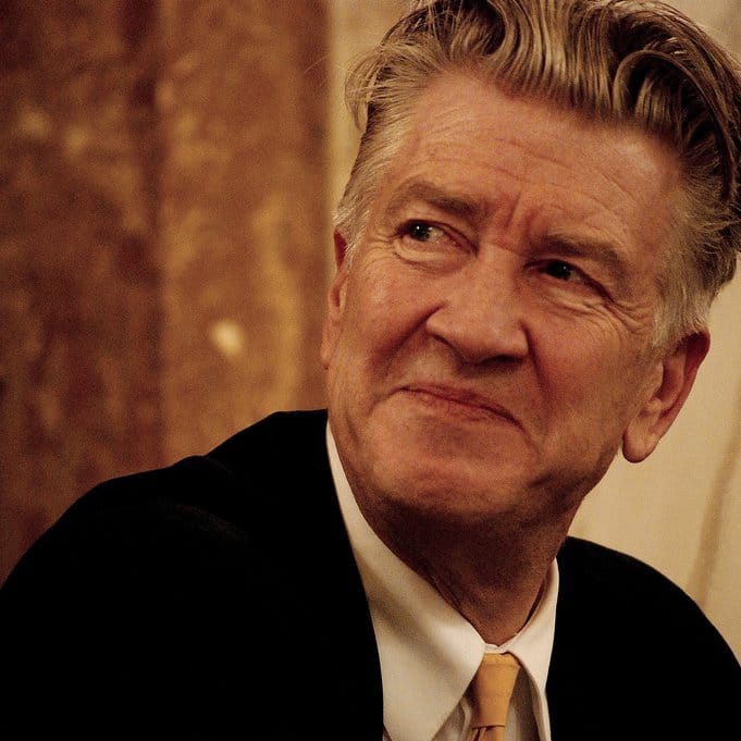 A smiling David Lynch in a soft black suit jacket, white shirt and honey-mustard-colored tie, looking at someone away from the camera