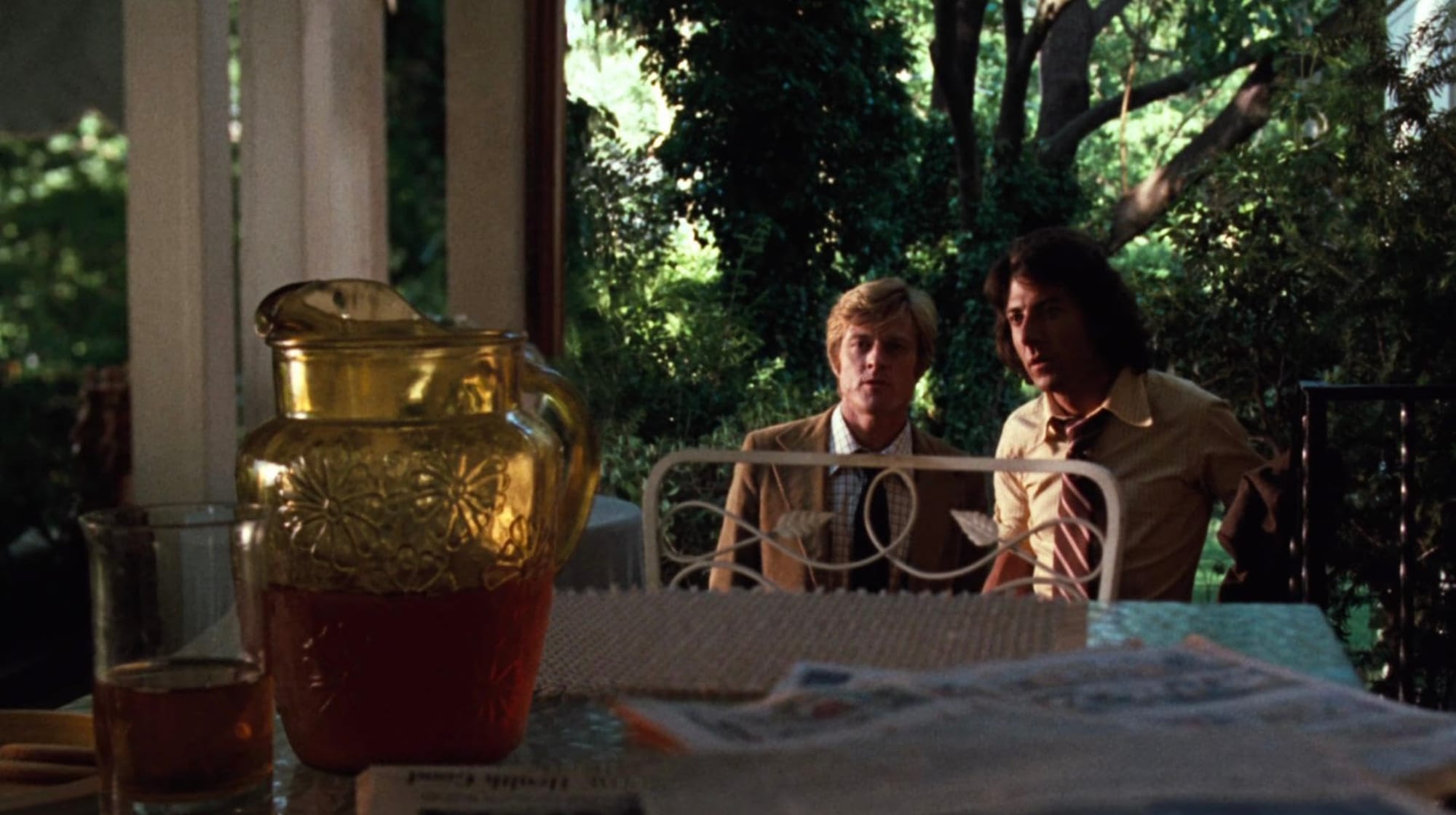Redford and Hoffman approaching a porch with a pitcher of iced tea in the foreground, trees and foliage behind them