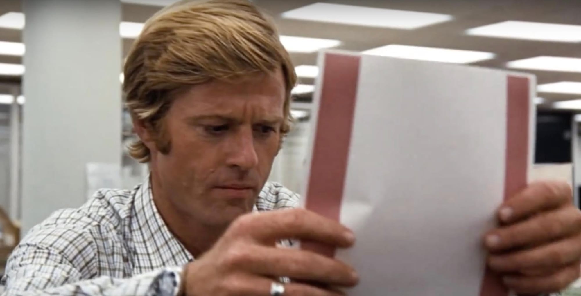 Robert Redford in a plaid shirt, reading intently, in the newsroom