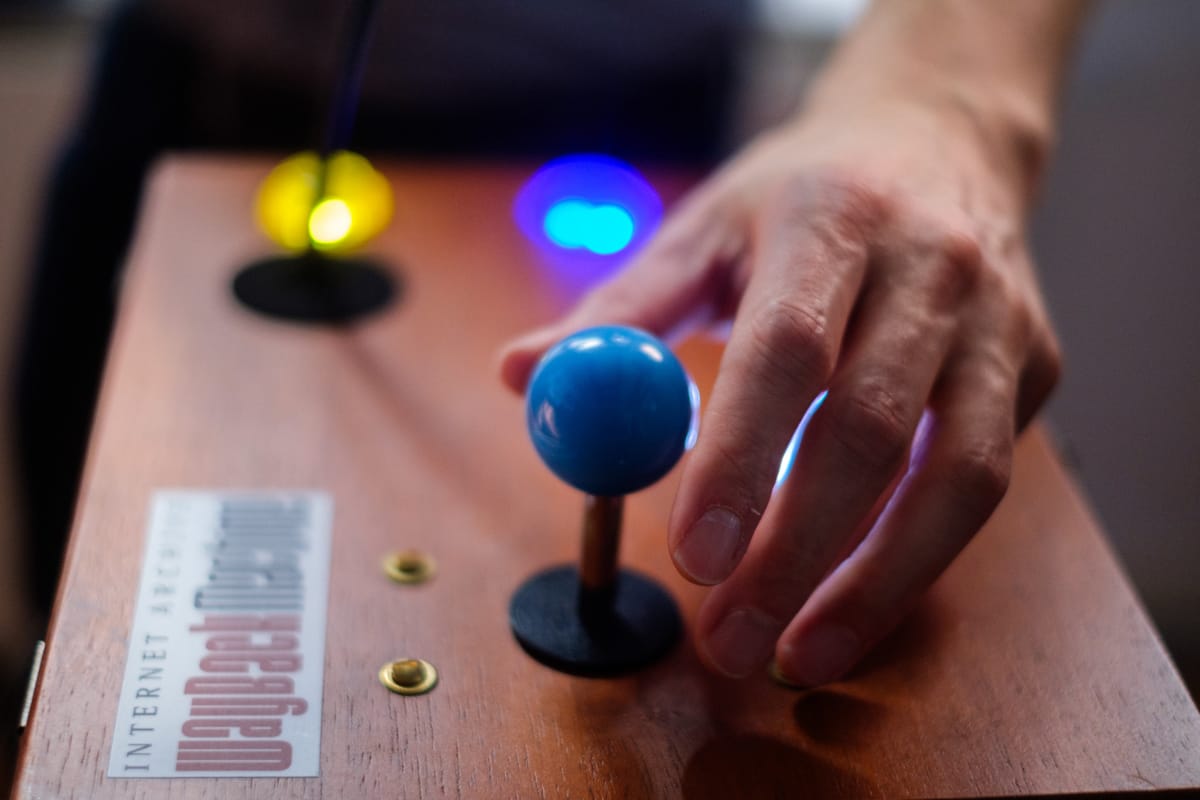 A hand reaches toward a blue joystick mounted on a wooden box with colorful lights on one side and a label at the edge reading, 'INTERNET ARCHIVE Wayback Machine'