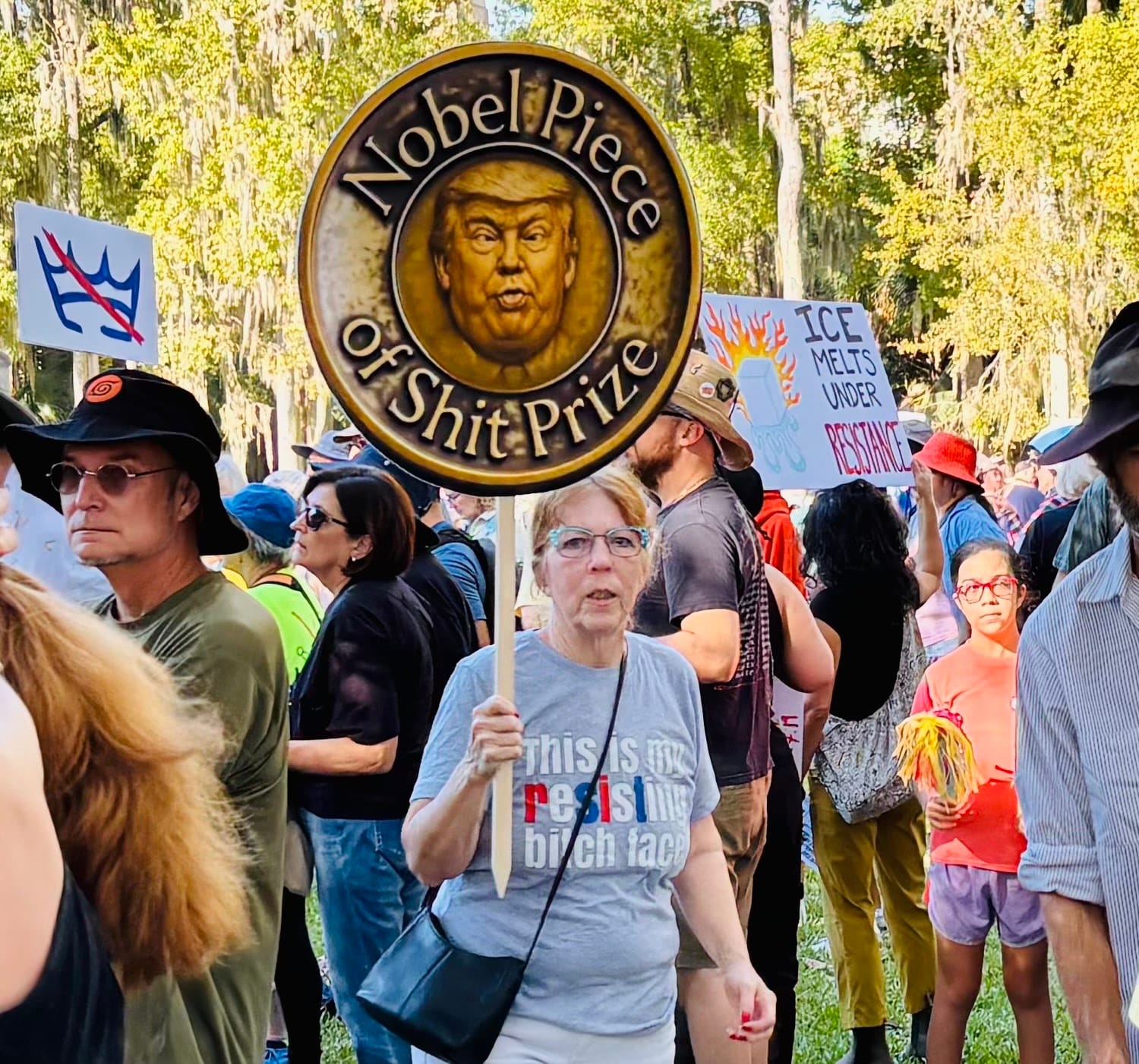 An older lady wearing a t-shirt reading, "This is my resisting bitch face" and holding a sign in the shape of a coin with Donald Trump's face and the legend, "Nobel Piece of Shit Prize"