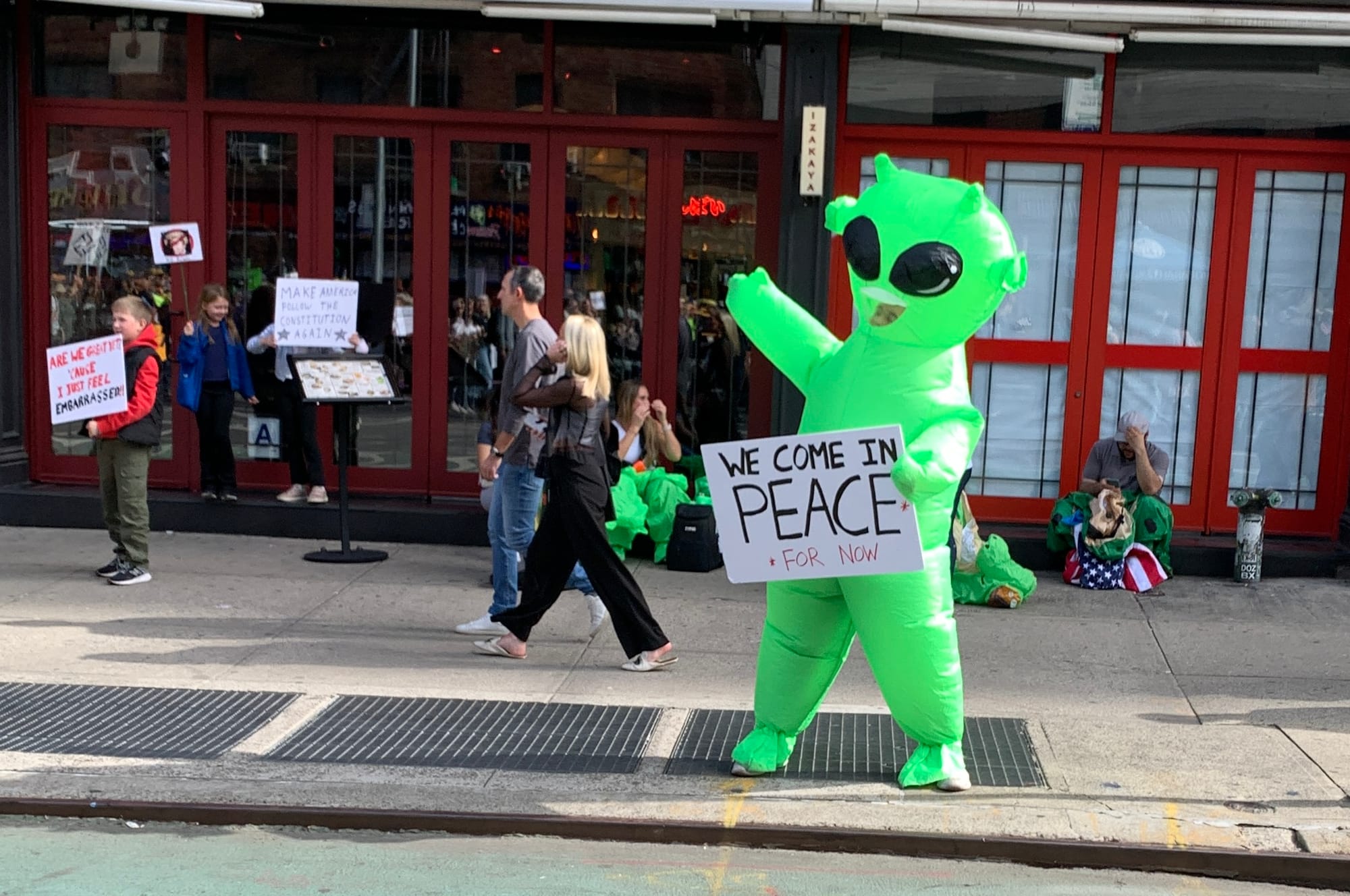 Inflatable green alien on the street holding sign: WE COME IN PEACE... FOR NOW