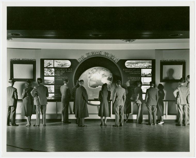 People standing in front of art deco-looking KODAK WORLD exhibit, a globe flanked by photos and signage. 