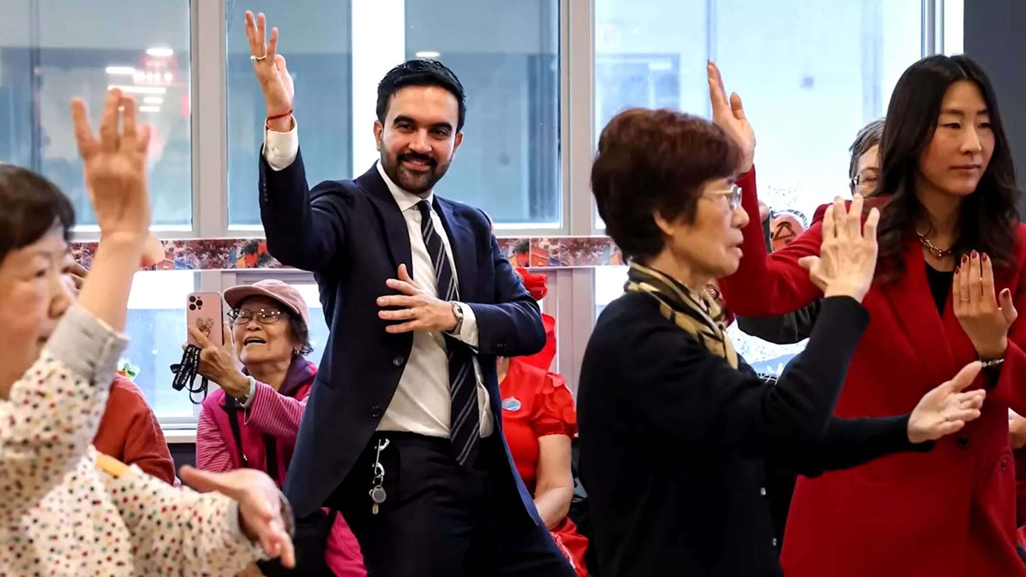 New York City mayor elect Zohran Mamdani, in suit and tie, practicing Tai Chi with a group of ladies before tall, sunlit windows.