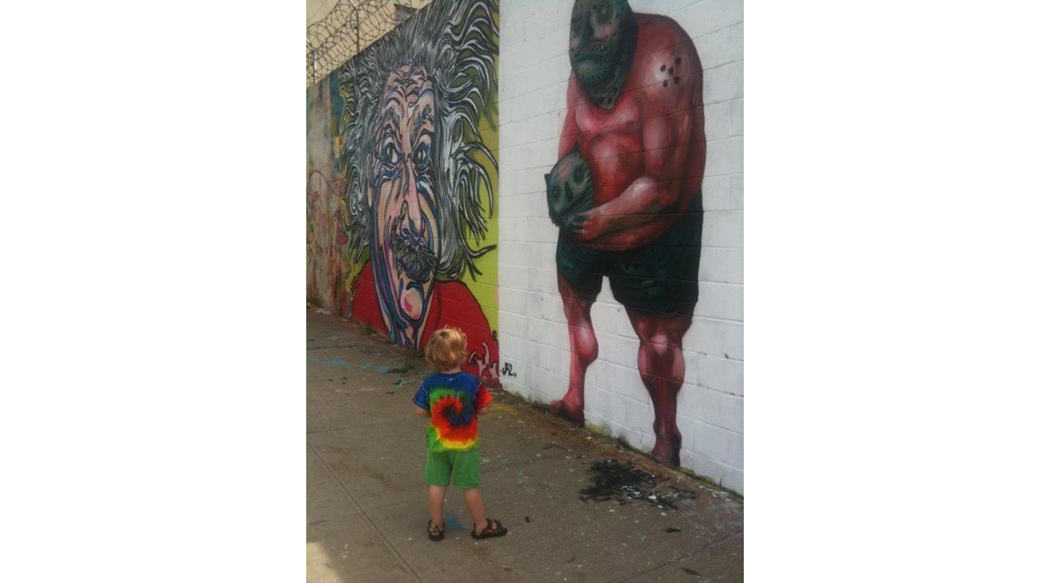 Small child wearing colorful tie dye shirt, in an alley regarding a white-painted brick wall featuring an eight-foot high painting of an ogre